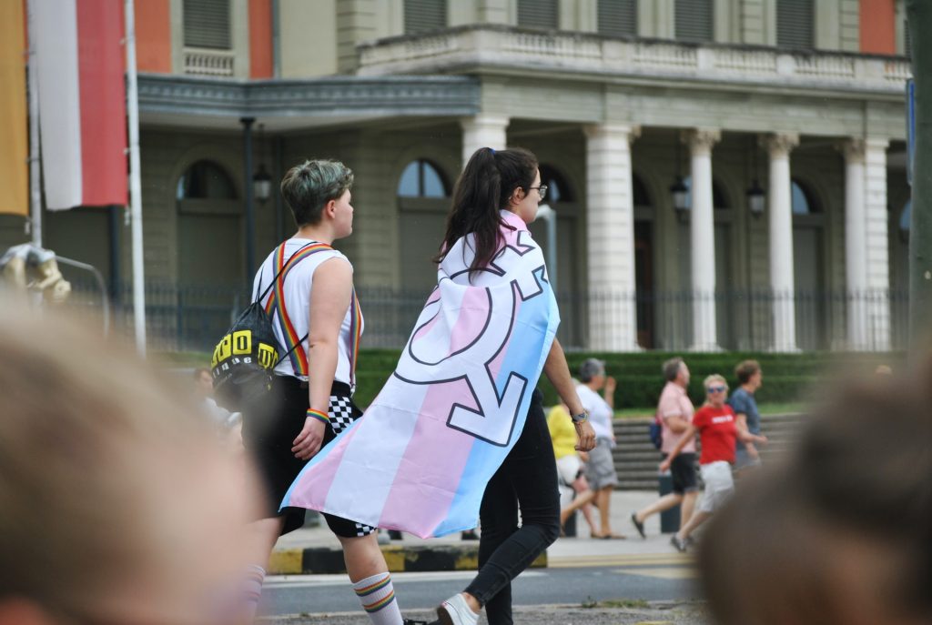 Two people walk together, one wears rainbow accessories, the other has a transgender flag as a cape